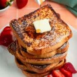Stack of brioche French toast slices topped with butter. Maple syrup is drizzling on top and strawberries are next to it on a white plate.