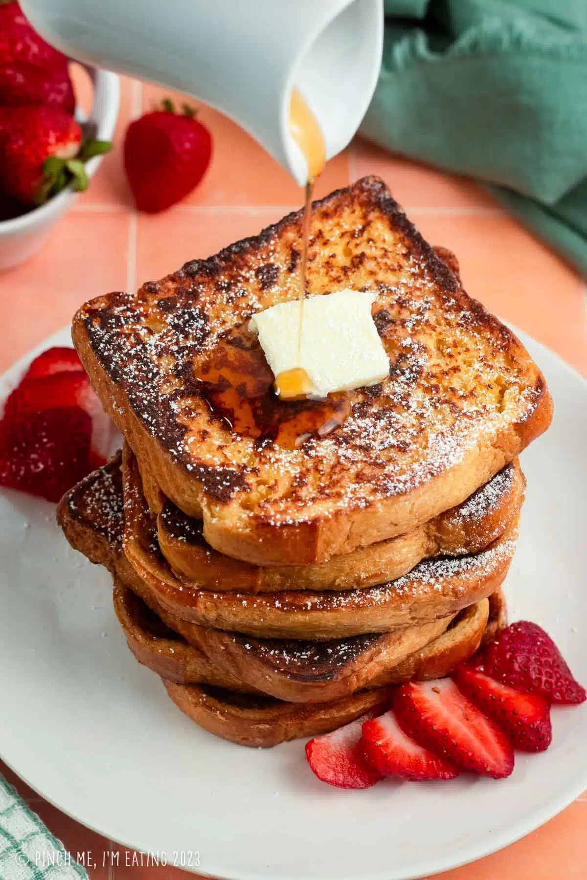 Stack of brioche French toast slices topped with butter. Maple syrup is drizzling on top and strawberries are next to it on a white plate.