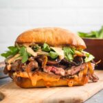 Grilled ribeye steak sandwich on ciabatta roll with roasted red pepper aioli, caramelized onions and mushrooms, and arugula on a cutting board.