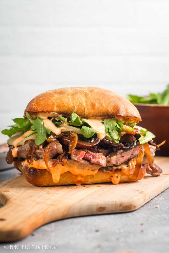 Grilled ribeye steak sandwich on ciabatta roll with roasted red pepper aioli, caramelized onions and mushrooms, and arugula on a cutting board.