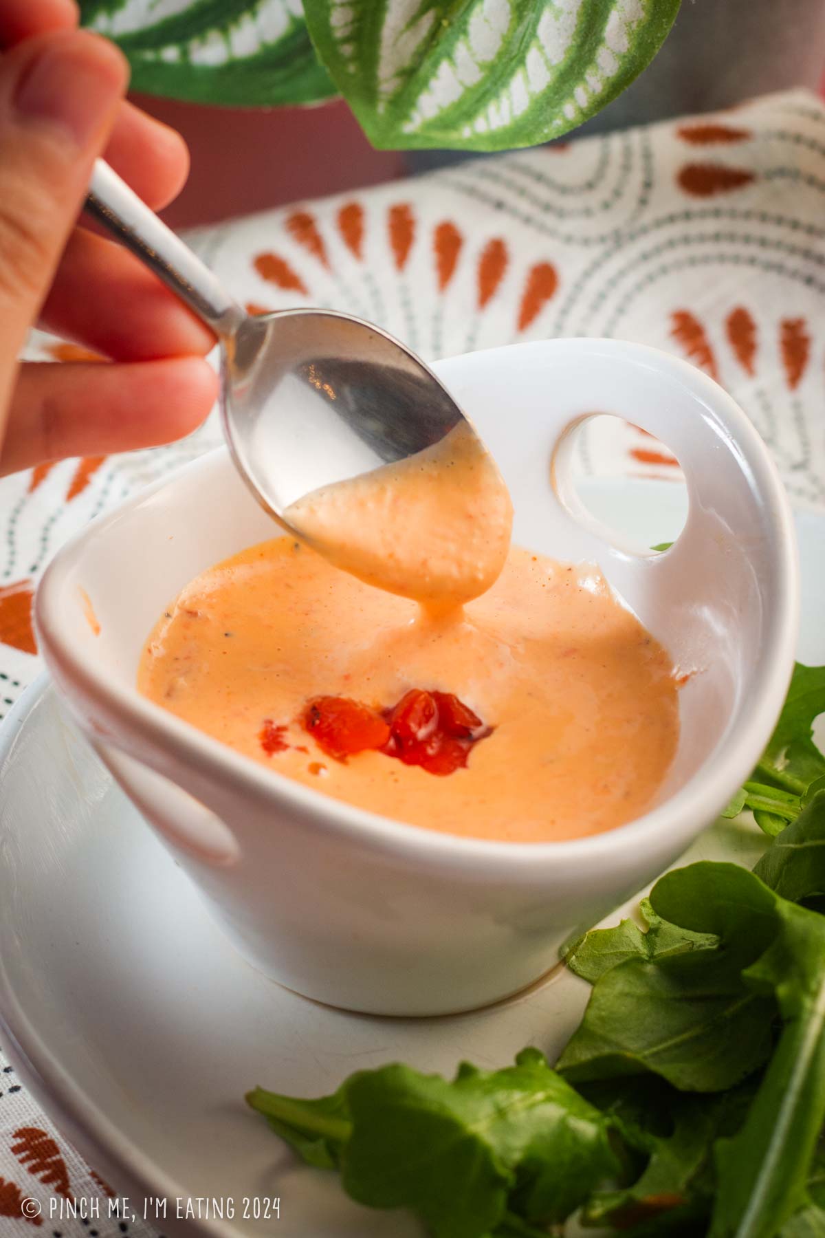 A spoon demonstrating the thick creamy consistency of roasted red bell pepper garlic aioli in a white bowl.