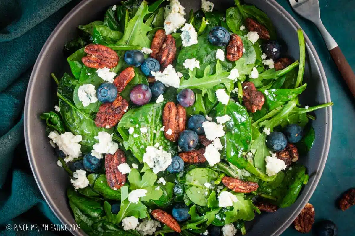Salad with spinach and arugula topped with blueberries, blue cheese, and candied pecans in a gray bowl.