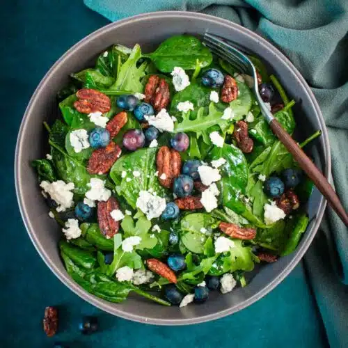 Spinach and arugula salad topped with blueberries, blue cheese, and pecans in a gray bowl.