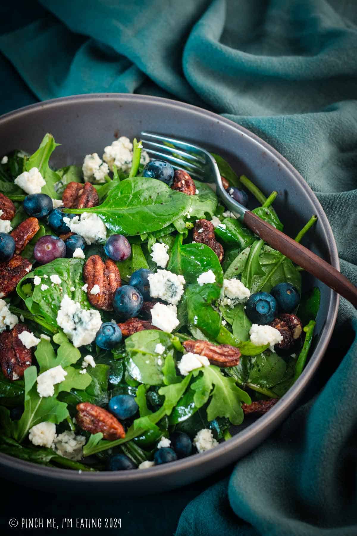 Arugula spinach salad topped with blueberries, blue cheese, pecans, and balsamic vinaigrette in a gray bowl.