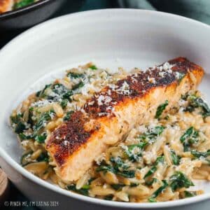 Blackened salmon fillet over bed of orzo and spinach in a white pasta dish, garnished with parmesan cheese.