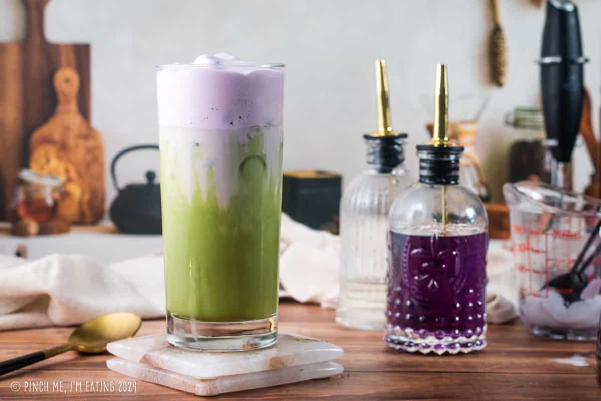 Homemade purple and green iced lavender cream oatmilk matcha latte in a tall glass next to bottles of simple syrup and lavender syrup.