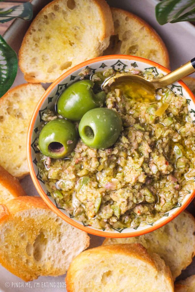 Green olive tapenade in a small bowl surrounded by crostini.