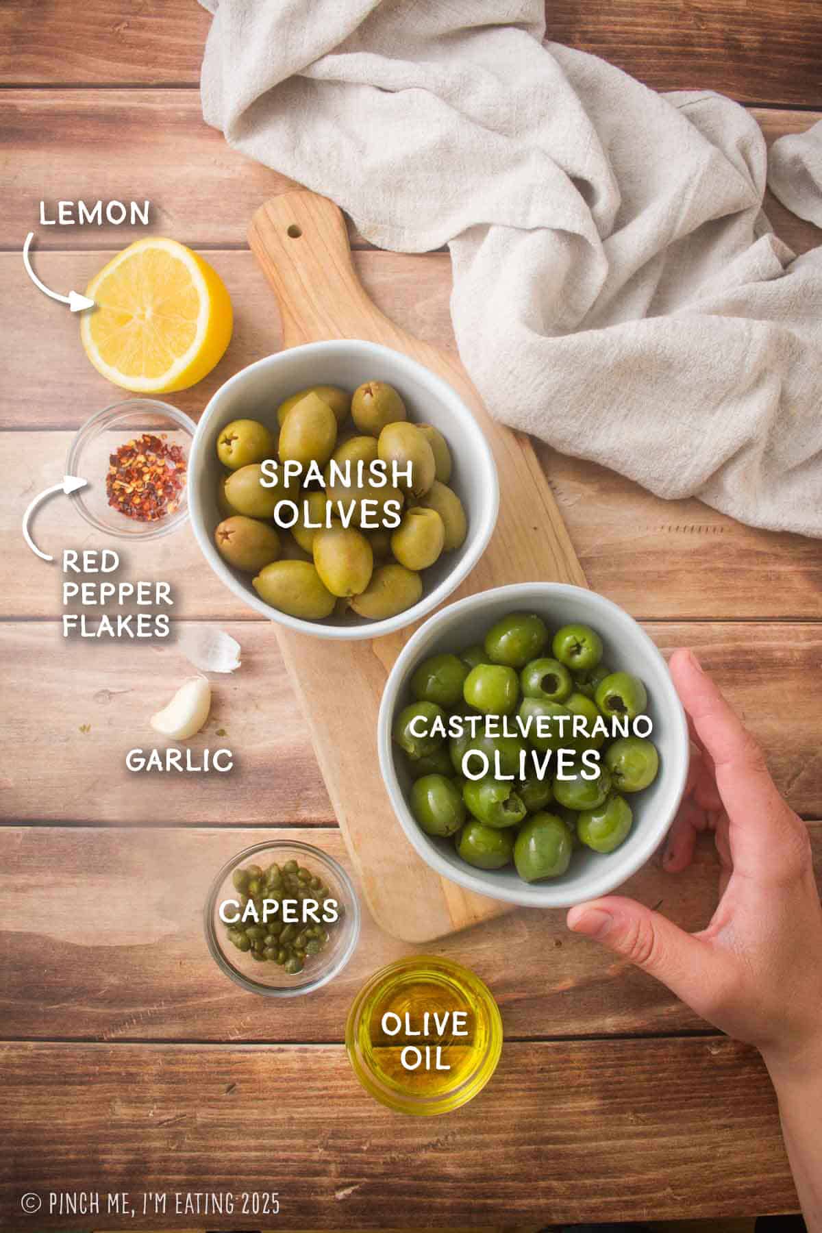 Labeled ingredient shot showing two types of green olives, capers, lemon, red pepper flakes, capers, a clove of garlic, and olive oil.
