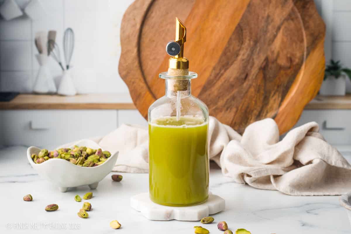 Green syrup in a bottle with pistachios around it on a marble countertop.