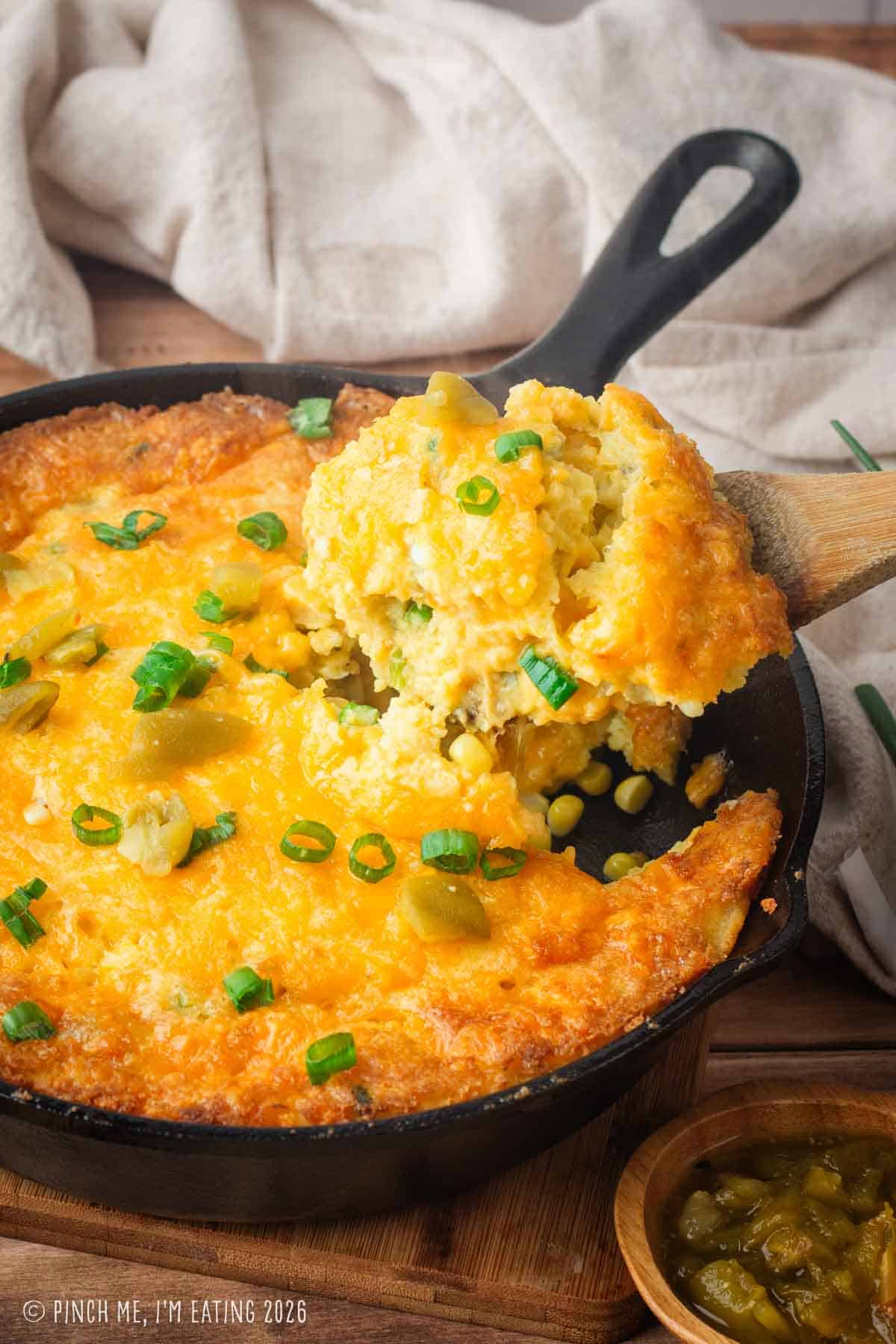 A vertical shot of a wooden spatula lifting a large scoop of custardy green chile corn pudding from a cast-iron skillet, showcasing the creamy interior and crispy cheese crust.