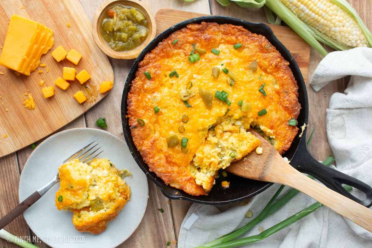 A homemade cast-iron skillet of Lewis Barbecue's famous Hatch green chile corn pudding recipe, featuring an overhead view of a crispy golden crust, melted cheese, green onions, and a scoop being lifted out with a wooden spatula.
