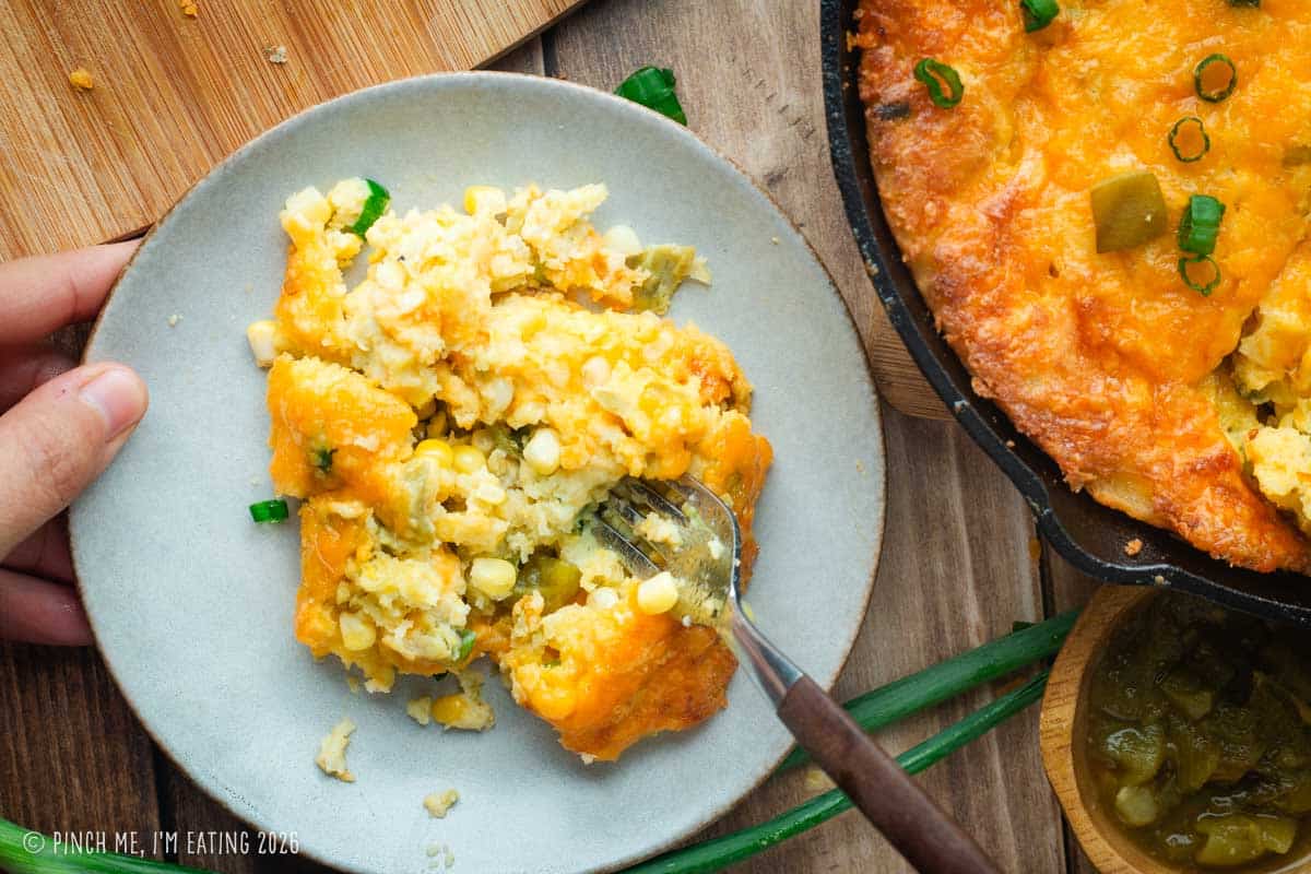 A close-up overhead shot of a serving of cheesy green chile corn pudding on a small plate, with a silver fork taking a bite to reveal the creamy, sweet corn texture.