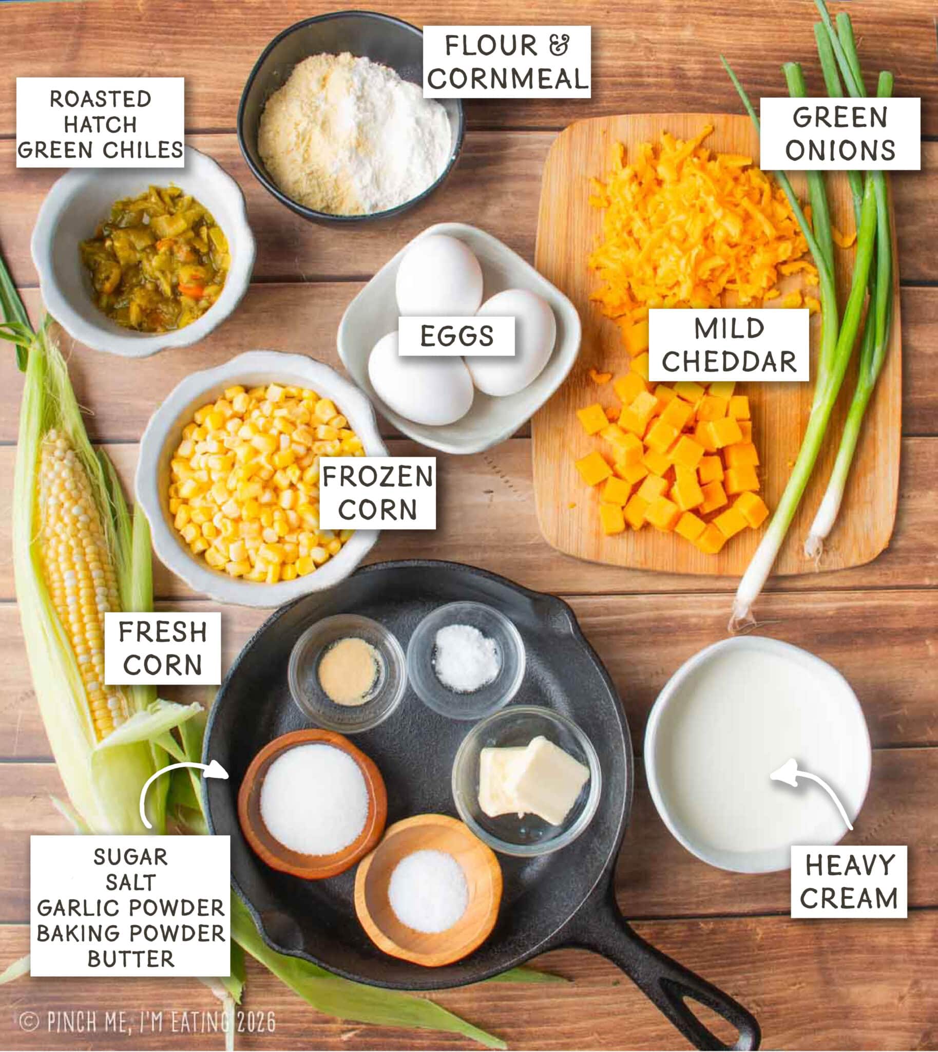 Flat lay overhead shot of ingredients for green chile corn pudding arranged on a wooden surface. Ingredients are labeled with text overlays and include an ear of fresh corn, bowls of frozen corn, roasted Hatch green chiles, eggs, flour and cornmeal, heavy cream, green onions, and a cutting board with shredded and cubed mild cheddar cheese. Small prep bowls containing sugar, salt, garlic powder, baking powder, and butter are arranged inside a black cast-iron skillet.