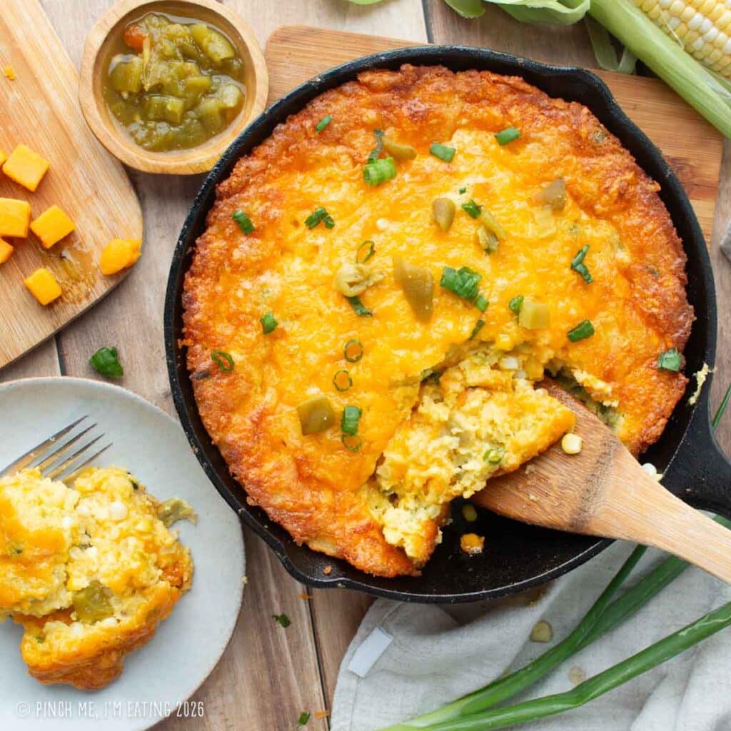 A homemade cast-iron skillet of Lewis Barbecue's famous Hatch green chile corn pudding recipe, featuring an overhead view of a crispy golden crust, melted cheese, green onions, and a scoop being lifted out with a wooden spatula.