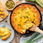 A homemade cast-iron skillet of Lewis Barbecue's famous Hatch green chile corn pudding recipe, featuring an overhead view of a crispy golden crust, melted cheese, green onions, and a scoop being lifted out with a wooden spatula.