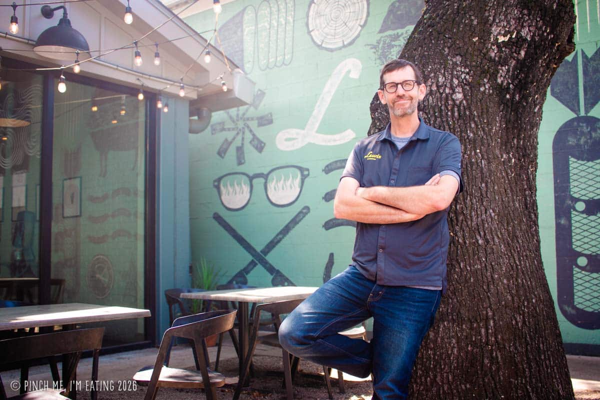 Chef and pitmaster John Lewis standing on the back patio of Lewis Barbecue, in Charleston, SC, leaning against a tree next to a painted green wall mural.