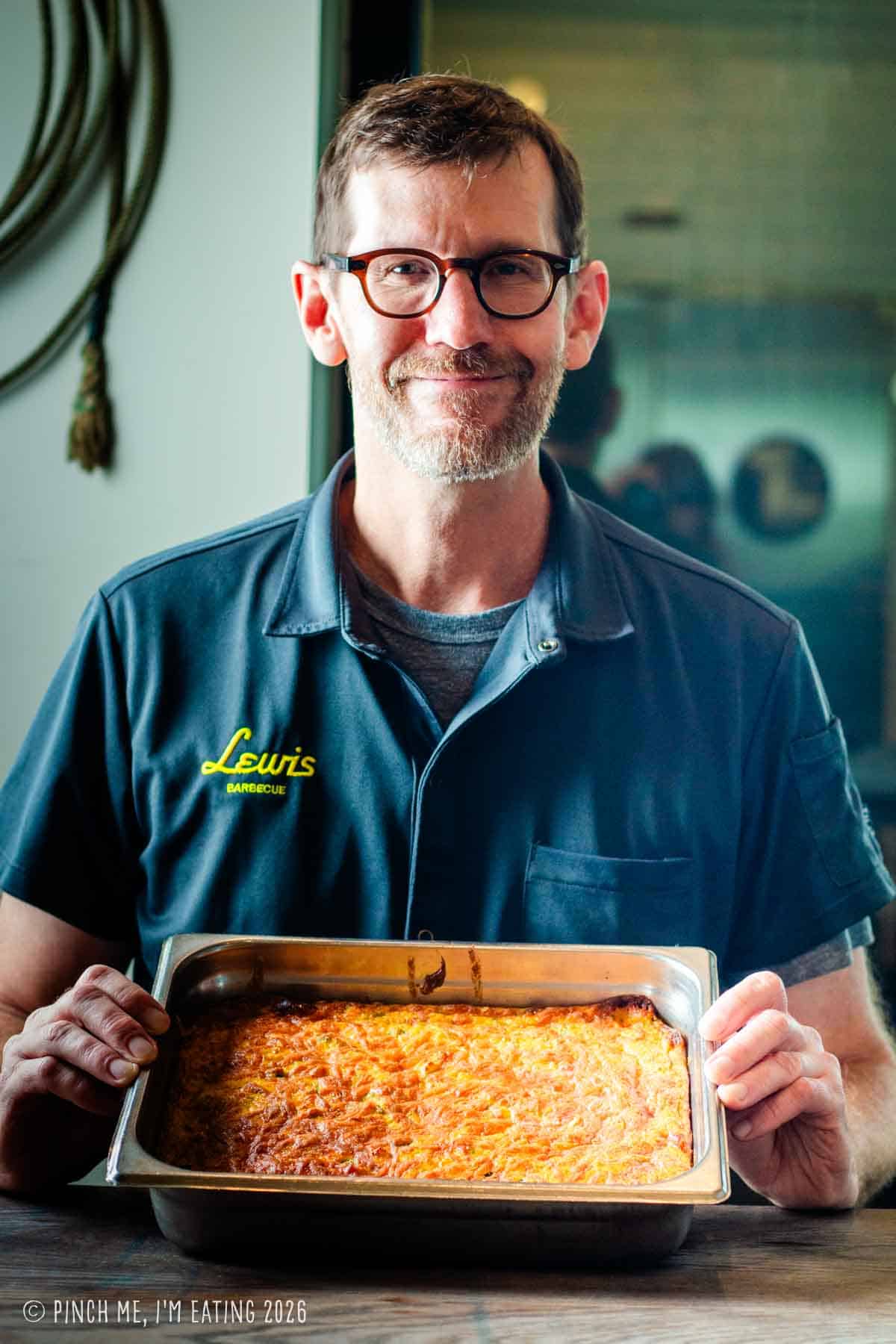 Pitmaster John Lewis smiling while holding a large, freshly baked catering pan of his famous Hatch green chile corn pudding.