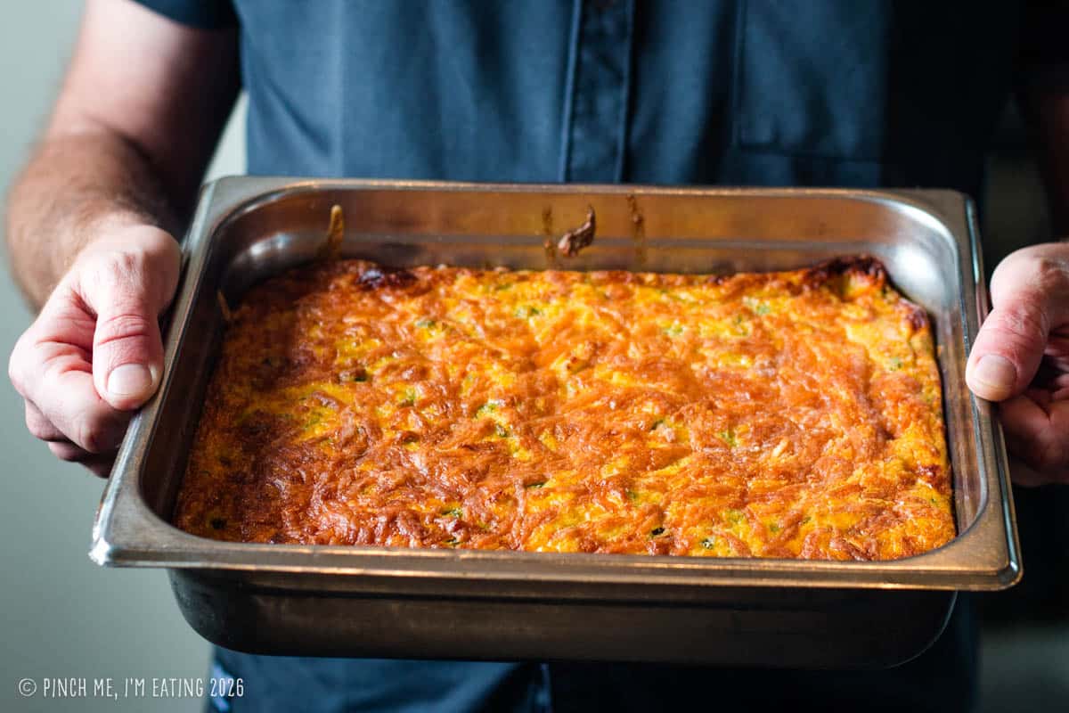 Close-up of pitmaster John Lewis's hands holding a large pan of baked green chile corn pudding, showcasing the golden-brown crispy-edged cheddar crust.