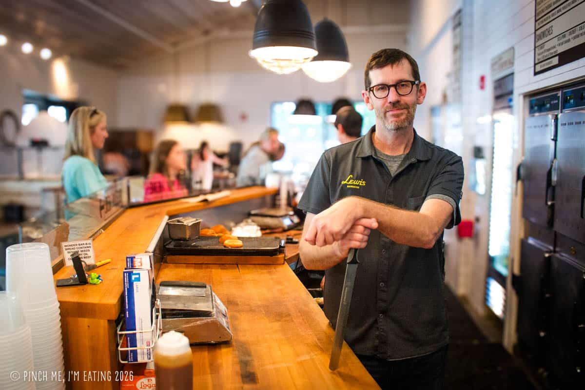 Pitmaster John Lewis standing at the wooden cutting counter inside Lewis Barbecue in Charleston, SC, resting his hands on a large meat-slicing knife.