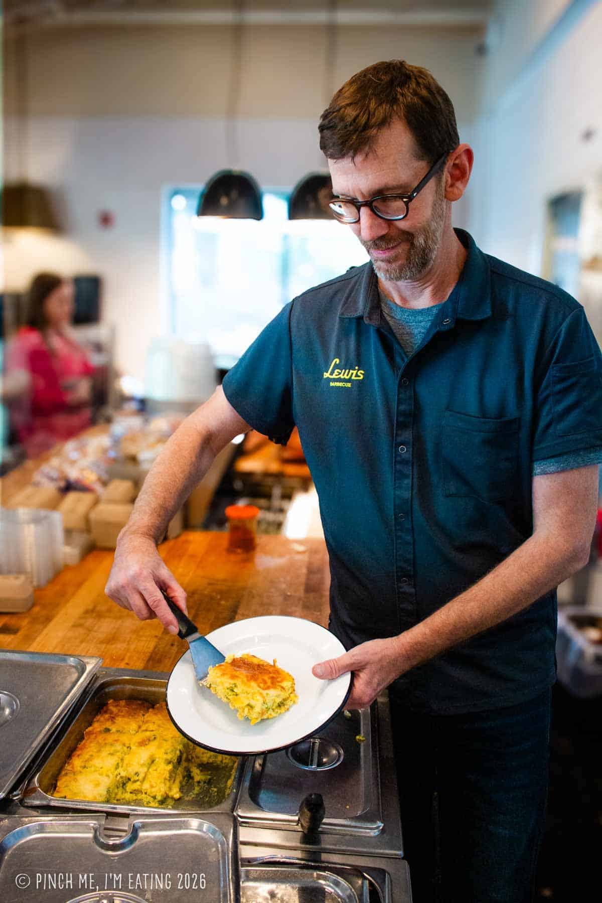 Pitmaster John Lewis of Lewis Barbecue serving a scoop of his signature green chile corn pudding onto a white plate inside the restaurant.