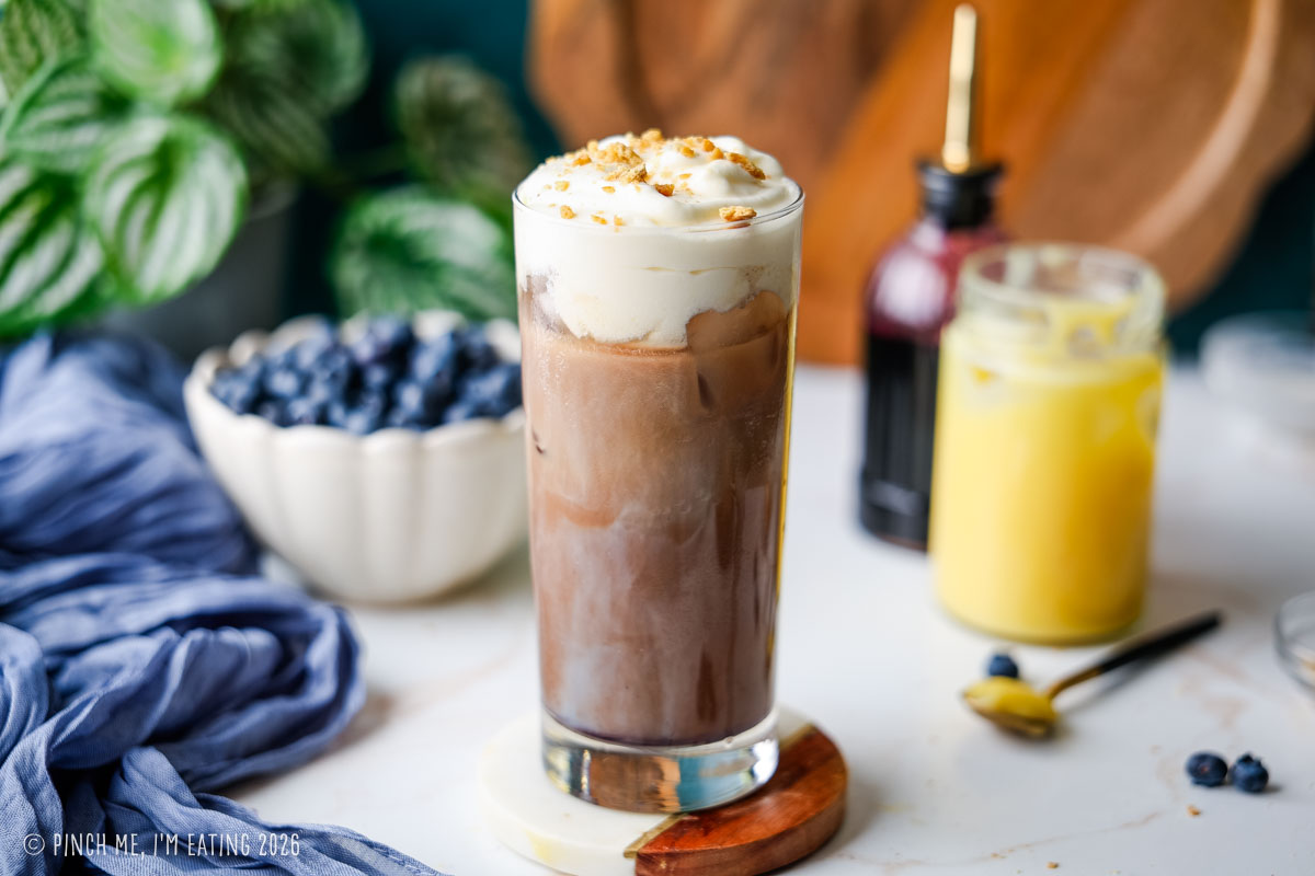 A iced blueberry latte topped with lemon curd cold foam and graham cracker crumbs in a tall glass in front of a bowl of blueberries, lemon curd, and blueberry syrup.